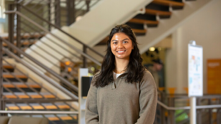 Grayson Nottage stands beneath the staircase in PLU's Anderson University Center. She is smiling and wearing a light brown sweatshirt.