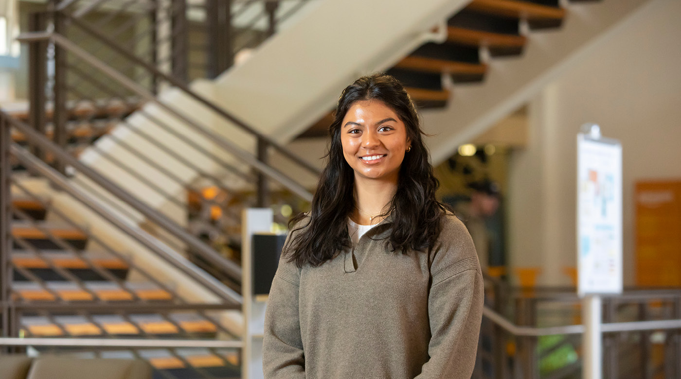 Grayson Nottage stands beneath the staircase in PLU's Anderson University Center. She is smiling and wearing a light brown sweatshirt.