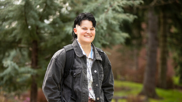 Allison Sheflo stands on campus with evergreen trees behind her. She's wearing a black denim jacket and smiling into the camera.