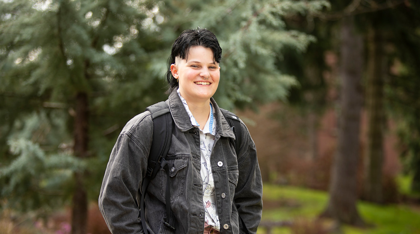 Allison Sheflo stands on campus with evergreen trees behind her. She's wearing a black denim jacket and smiling into the camera.