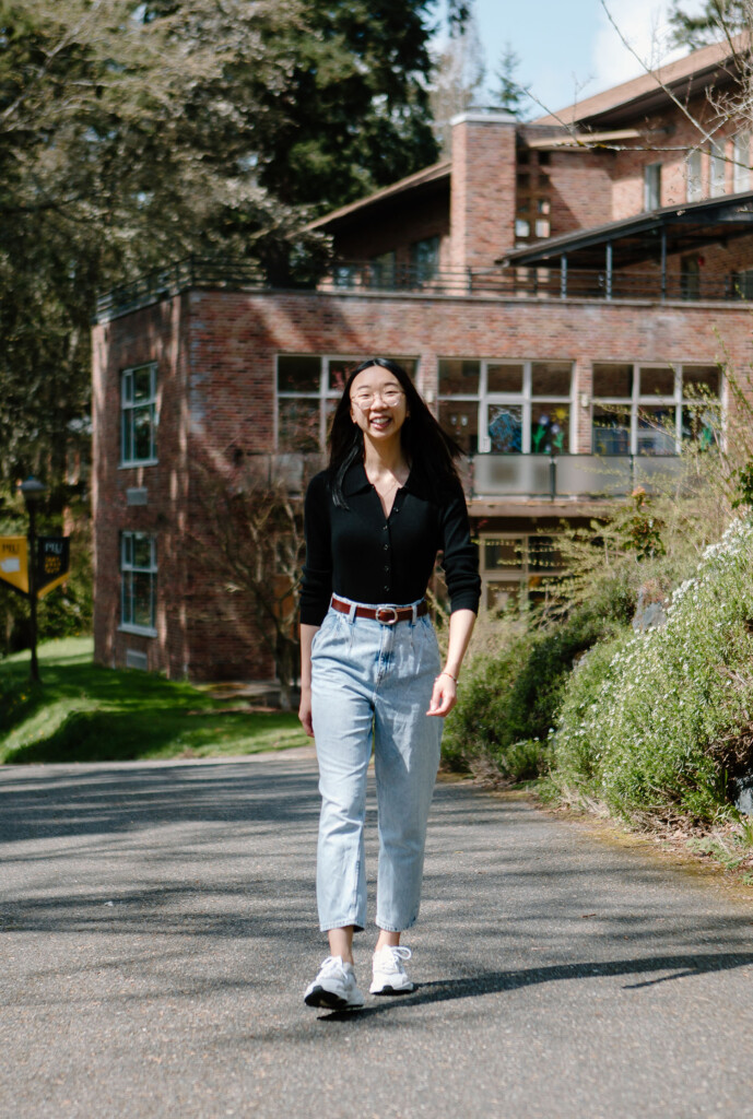 Tiffany Wong walking on campus at PLU. She is smiling and wearing a black blouse with blue denim pants.