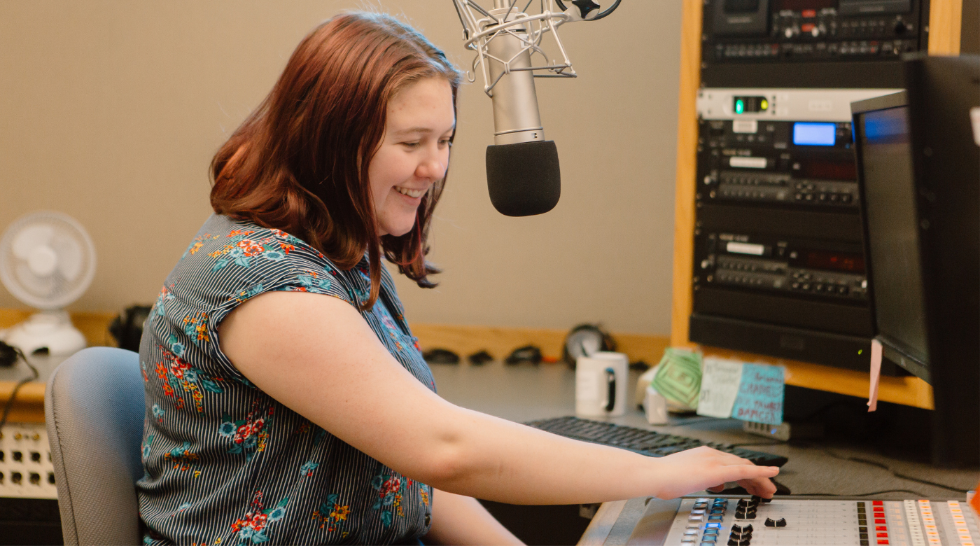 PLU student sits at a soundboard in a recording studio and talks into a mic. They are wearing a flower short sleeve shirt and have shoulder length red hair.