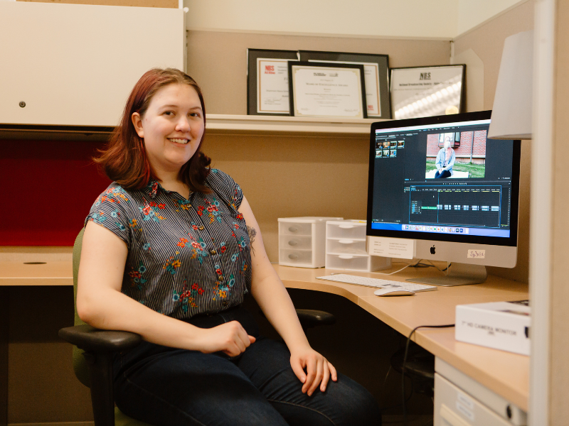 PLU student sits at a desk in front of a computer working on video editing software. They are wearing a flower short sleeved top and blue jeans. They are smiling into the camera.