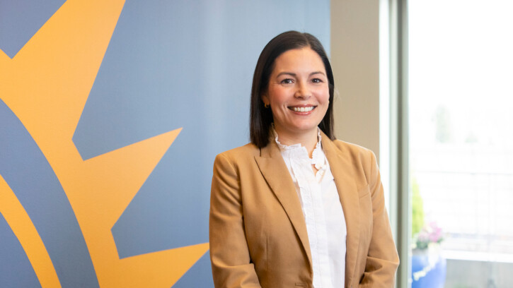 Lange smiles while standing in the lobby of her office. She is wearing a tan blazer over a white shirt and a blue and gold wall is behind her.