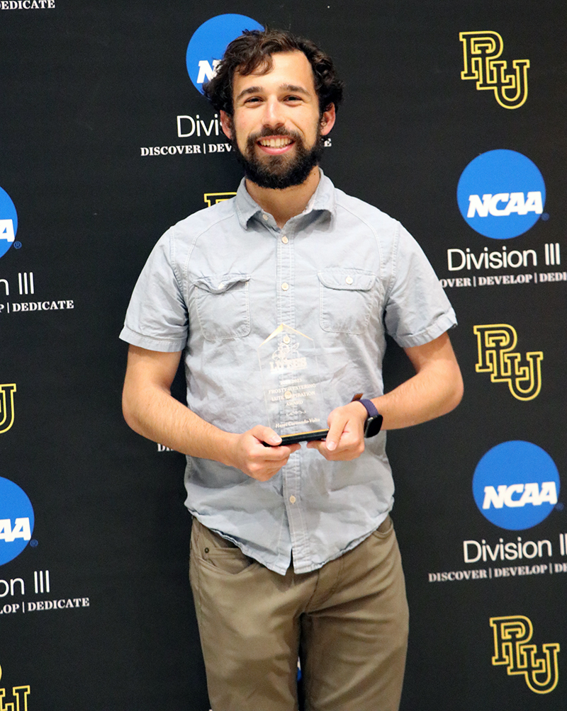 PLU student Henri Coronado-Volta holds an award while smiling into the camera. He is wearing a short sleeve gray button-down shirt and khaki pants. He is standing in front of a PLU and NCAA banner.