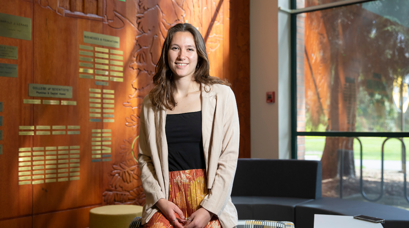 PLU student, wearing a black shirt, cream cardigan sweater, and orange and yellow pants, smiles into the camera while standing in front of a wooden wall and window.