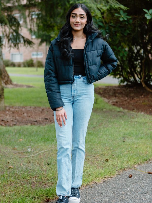 PLU student stands in front of a tree smiling into the camera. Wearing a blue jacket, black shirt, jeans, and blue sneakers.