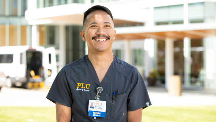PLU nursing student wearing dark gray scrubs and a name tag faces the camera and looks up and smiles. The student is standing in front of a hospital. It is sunny outside.