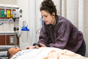 A nursing student is working with a mannequin that is laying in a bed. The student is wearing a purple sweatshirt and is checking the vitals of the mannequin.