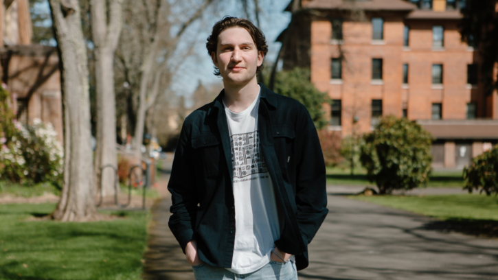 PLU student wearing a black jacket over a white t shirt looks into the camera. They are standing in front of trees and a brick building.