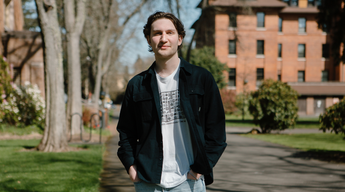 PLU student wearing a black jacket over a white t shirt looks into the camera. They are standing in front of trees and a brick building.