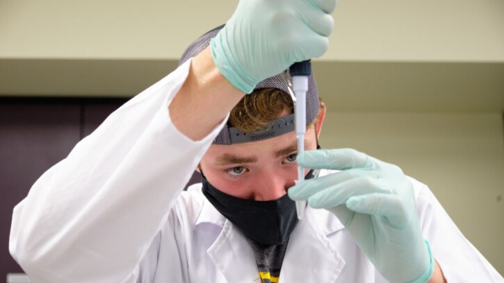 A student wearing a black mask, a blue baseball cap turned backward, and a white lab coat holds a pipette in front of their face as they practice adding liquid to a pipette.