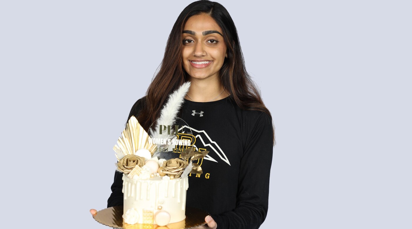 PLU students hold a highly decorated cake while smiling into the camera. The student is a PLU rowing sweatshirt.