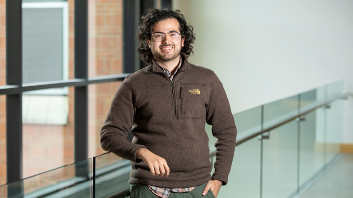 Chris Holland smiles into the camera while leaning on a glass railing. He is wearing a brown sweatshirt with windows in the background.