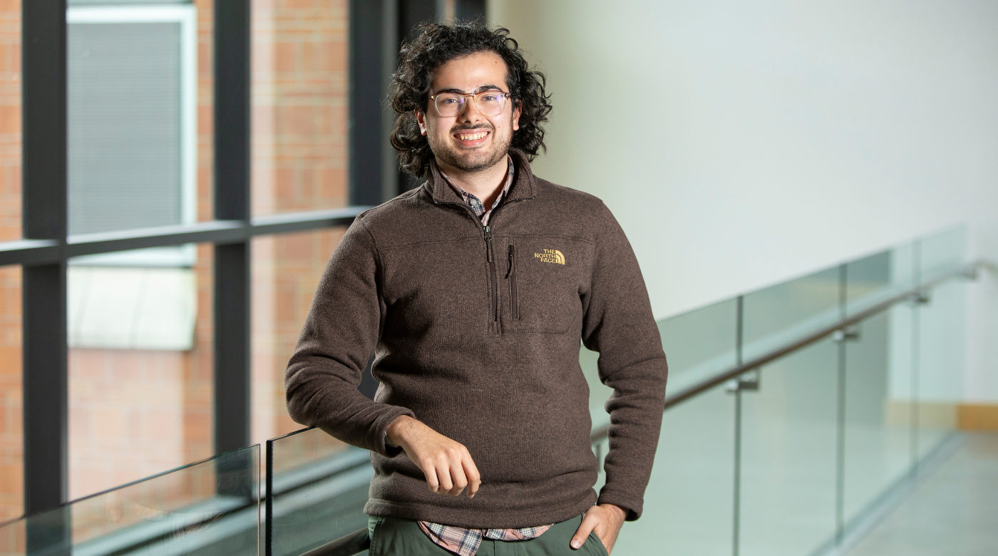 Chris Holland smiles into the camera while leaning on a glass railing. He is wearing a brown sweatshirt with windows in the background.