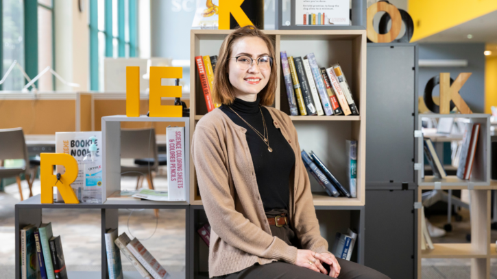 Emily Struck sitting in the library in front of a stack of books, smiling into the camera. She has glasses and is wearing a black shirt with a brown sweater. Her hands are in her lap.