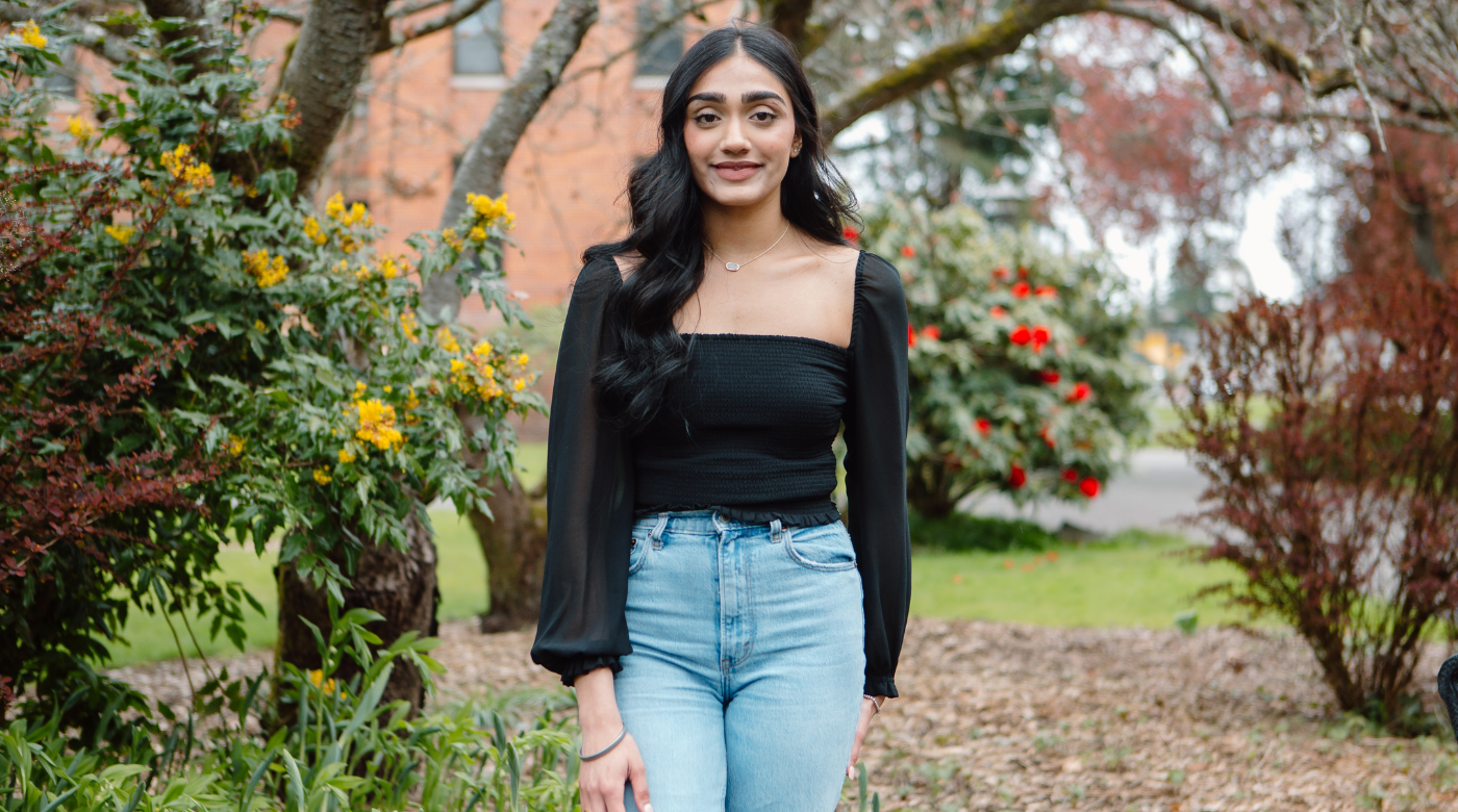 PLU student wearing a black top and jeans smiles into the camera. They are surrounded by flowers and trees.