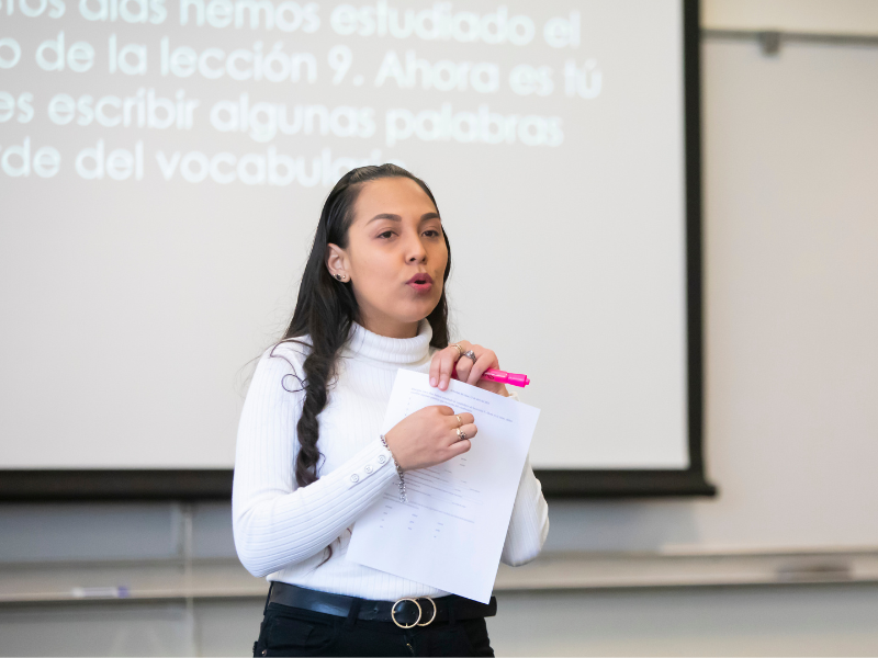 PLU Professor is wearing a white turtle neck and black pants with belt. They are holding a paper and pink highlighter. They are standing in front of a projector screen with Spanish words on it.