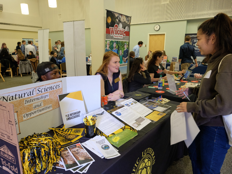 Students attend a internship and career fair. Two PLU students are sitting at a table covered with papers and Lute gear. They are looking at and talking to a third PLU student.