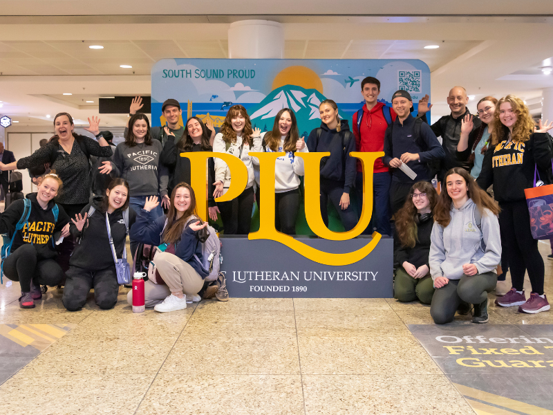 PLU students stand in front of a PLU sign at the SeaTac airport before they board a flight.