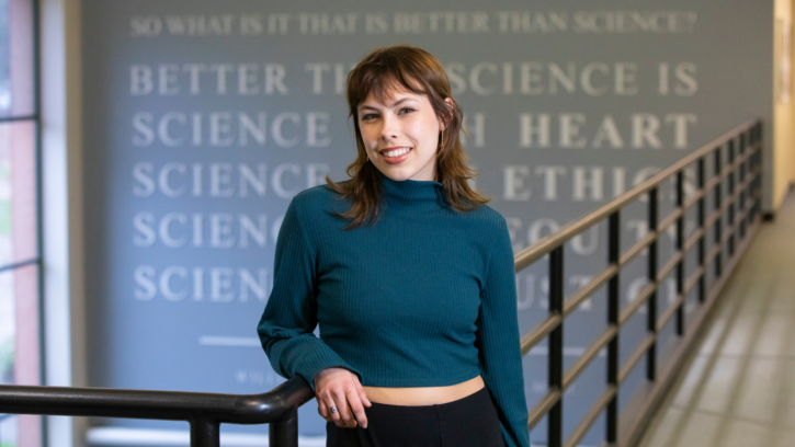 PLU student, wearing a dark blue turtle neck shirt and black pants, leans on a banister while smiling into the camera. They are standing in front of a quote about science on the back wall.