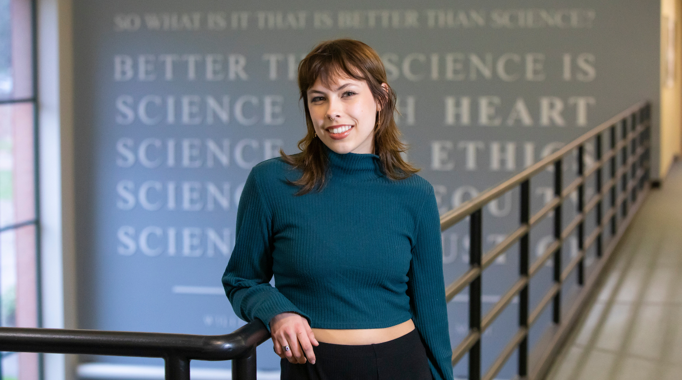 PLU student, wearing a dark blue turtle neck shirt and black pants, leans on a banister while smiling into the camera. They are standing in front of a quote about science on the back wall.