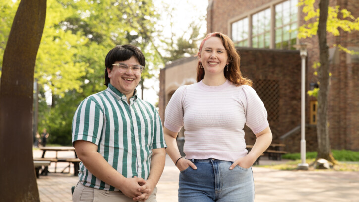 Two PLU students stand next to each other and smile into the camera. The student on the left is wearing a green striped shirt and the student on the right is wearing a pink short sleeved shirt. The photo is taken outside in front of a building on campus.