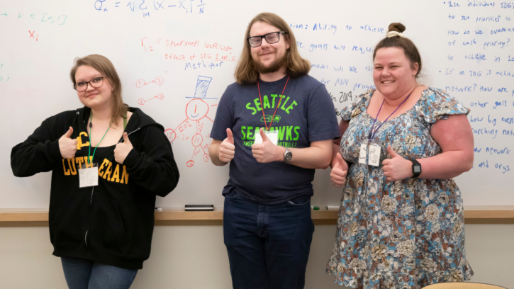 Three PLU students look into the camera and make a thumbs up sign with their hands. Behind them is a whiteboard with a math problem on it.