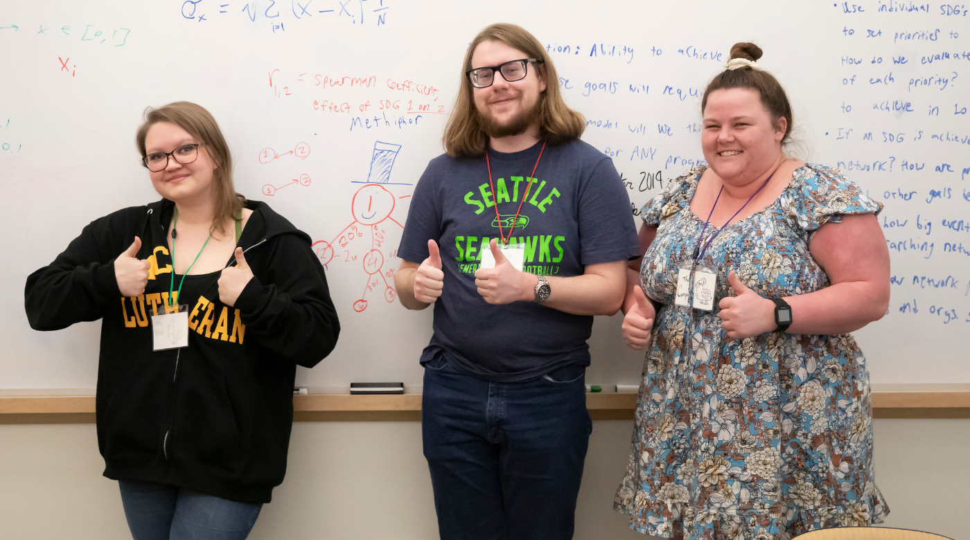 Three PLU students look into the camera and make a thumbs up sign with their hands. Behind them is a whiteboard with a math problem on it.