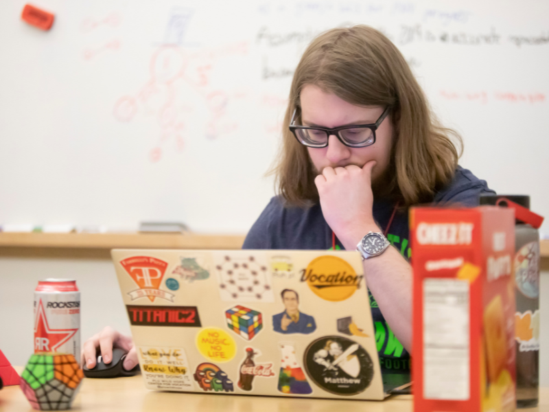 One student hold their hand to their chin while looking at a laptop covered in stickers.