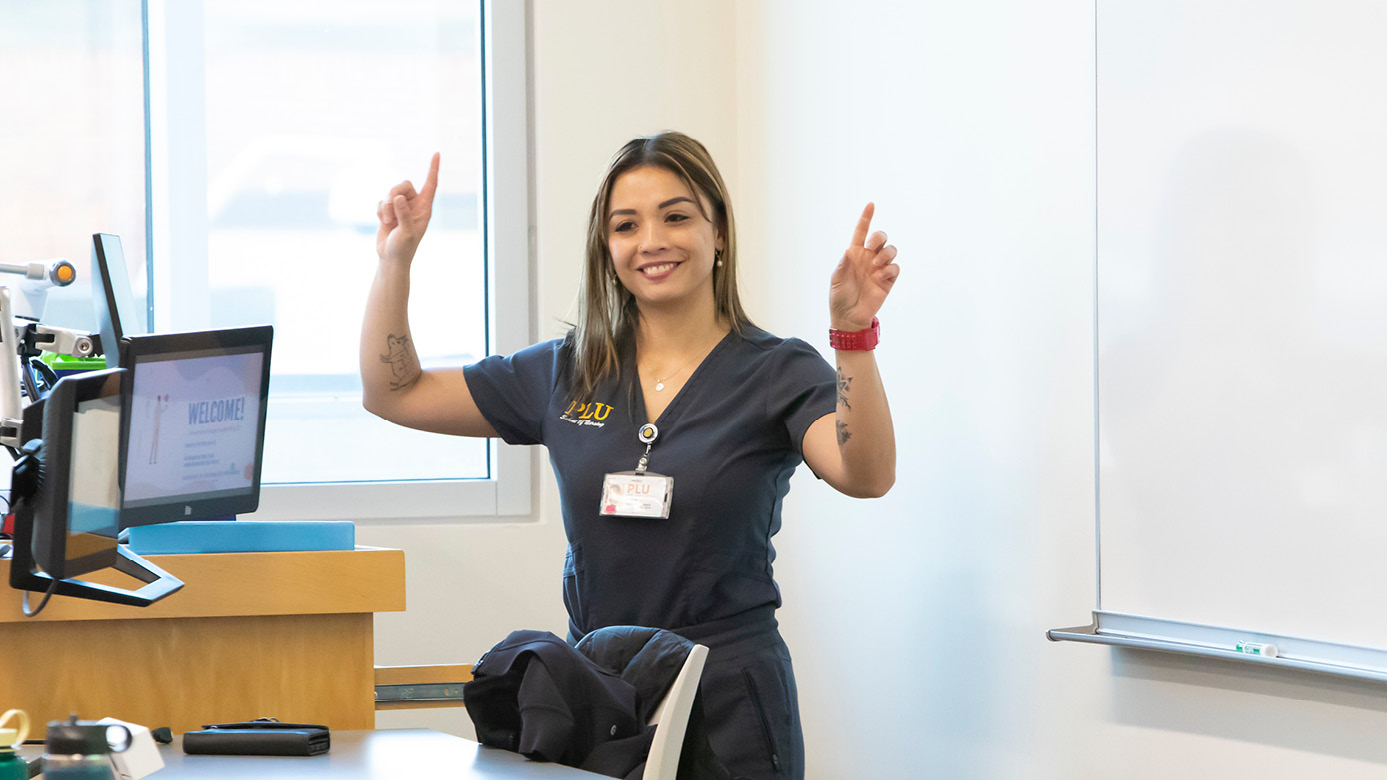 Maria Surla stands in front of a nursing classroom. She is smiling, wearing nursing scrubs, and gesturing up with both of her hands.
