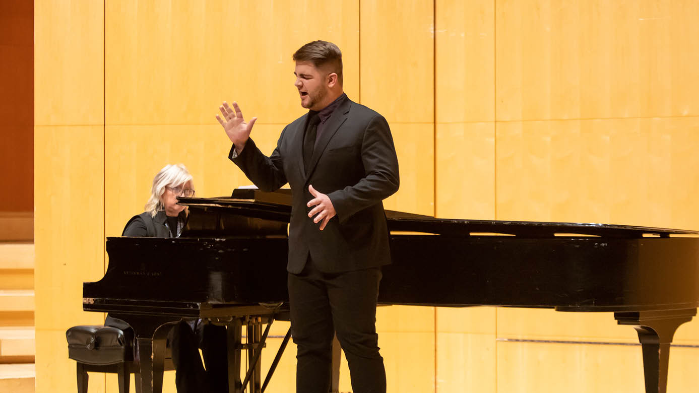 PLU student Jack Burrows lifts his right hand for effect as he sings on a PLU music stage. He is wearing a black shirt and black suit, and a piano player is behind him.