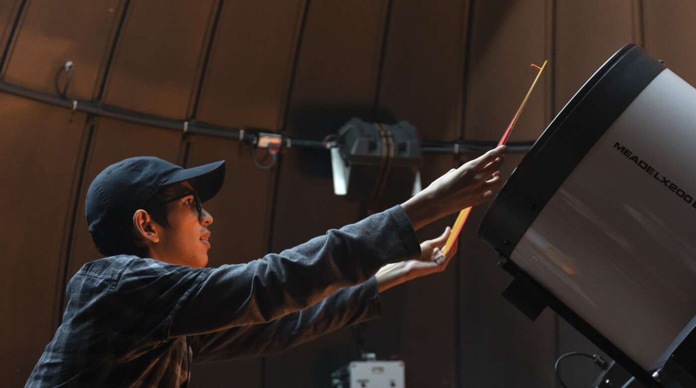 PLU students is standing on a ladder in front of a large telescope placing a mask over the front of the telescope.