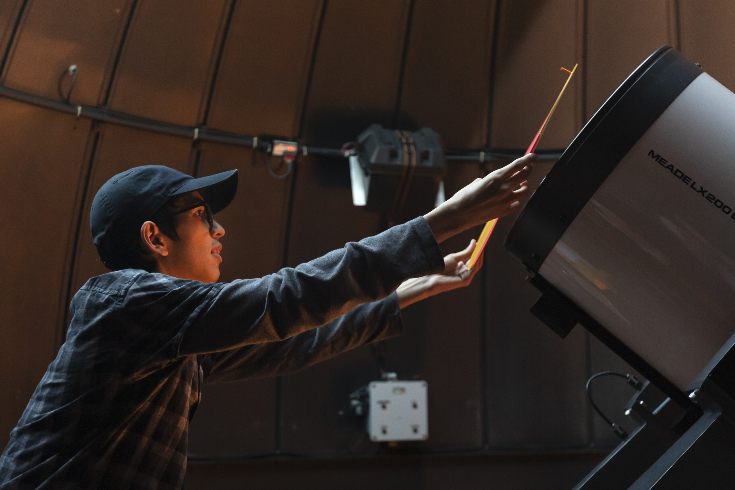 PLU students is standing on a ladder in front of a large telescope placing a mask over the front of the telescope.