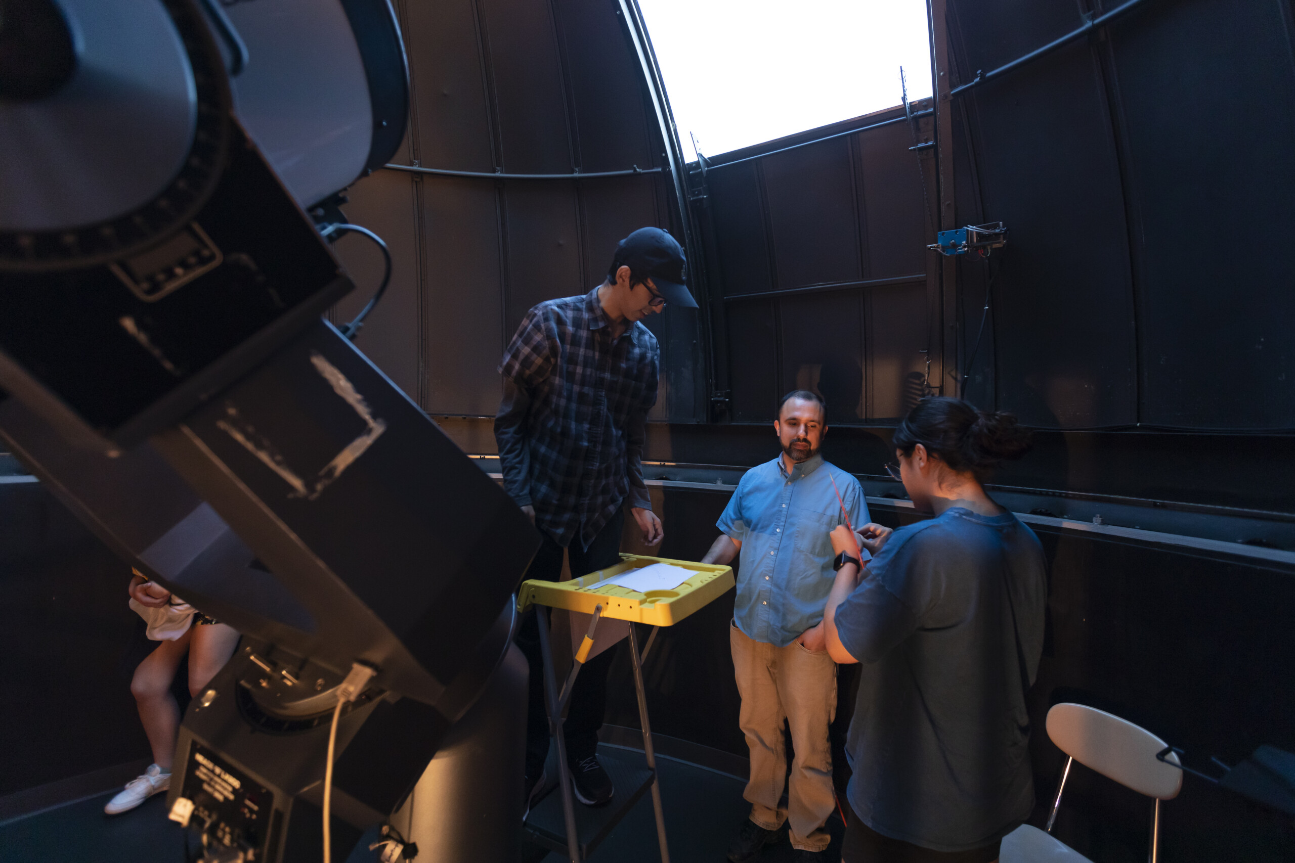 Two students and a professor stand in the observatory looking at the telescope.