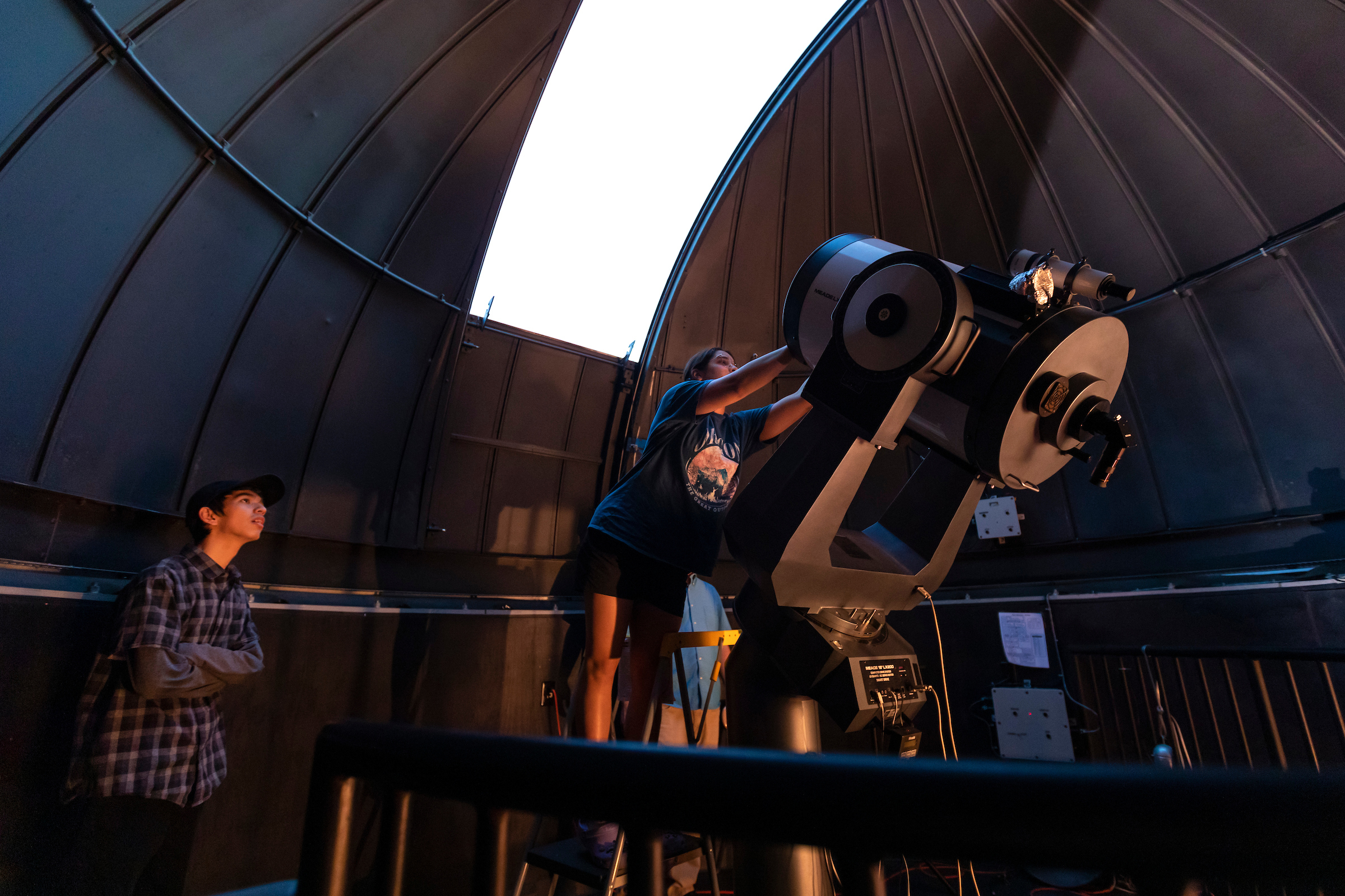 Student stand in front of large telescope. Another student watches with their arms folded.