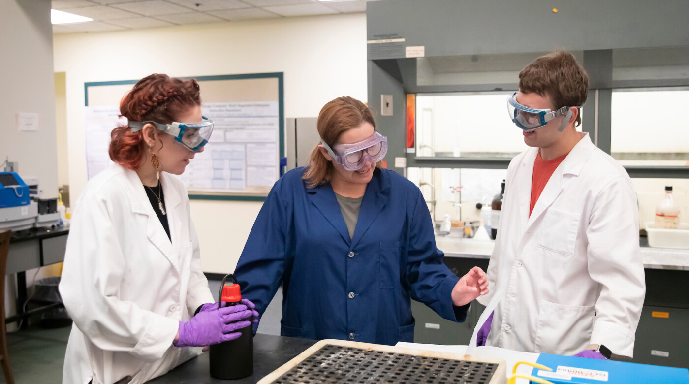 Two students talk with a professor as they lean over a chem lab table.