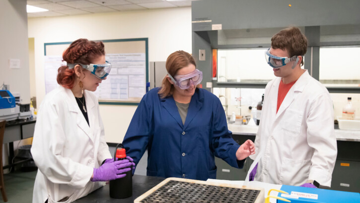 Two students talk with a professor as they lean over a chem lab table.