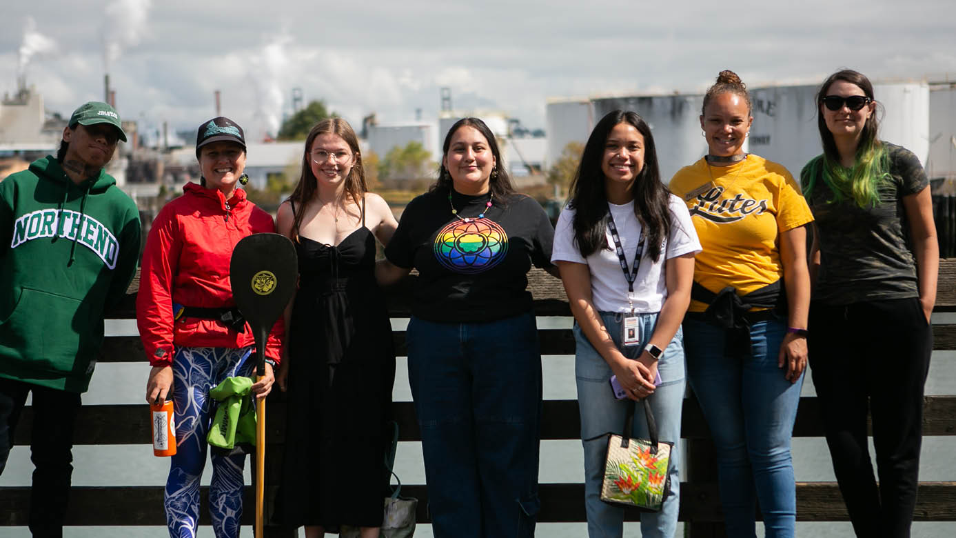 7 PLU students, staff and alumni stand in a row with commencement bay behind them.