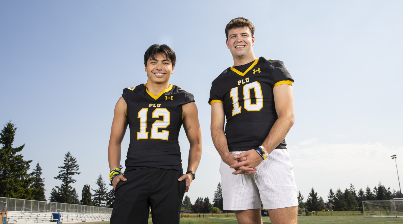 Two PLU football players stand in front of the practice field wearing their uniforms and smile into camera.