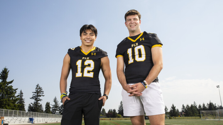 Two PLU football players stand in front of the practice field wearing their uniforms and smile into camera.