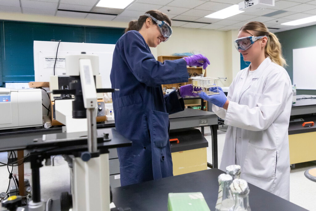 Professor wearing a blue lab coat works with a student wearing a white lab coat in a science lab.