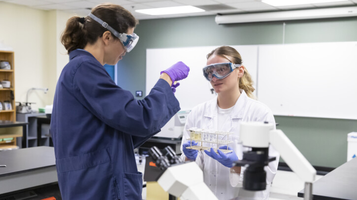 A professor in a blue lab coat looks at a sample in the lab with a student wearing a white lab coat.