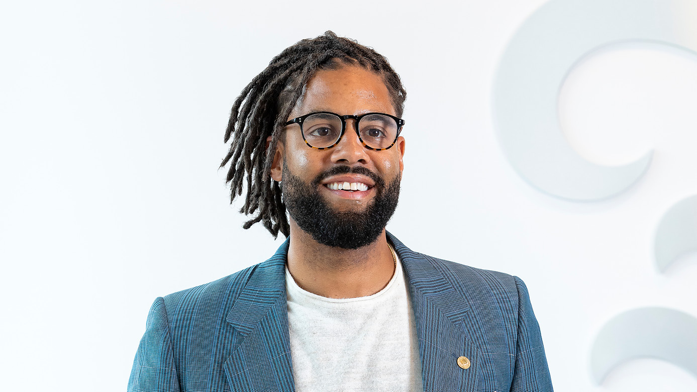 Aaron Bell smiles at the camera. There is a white wall behind him and he is wearing a blazer and glasses.