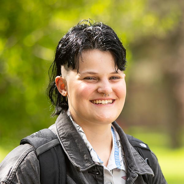 Allison Sheflo stands on campus with evergreen trees behind her. She's wearing a black denim jacket and smiling into the camera.