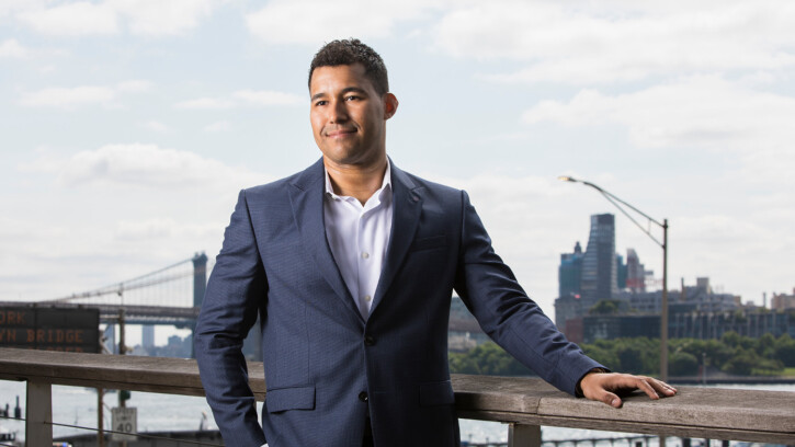 Dayton Campbell-Harris stands on a park deck with water and the Brooklyn skyline behind him. He's wearing a blue blazer and half-smiling.