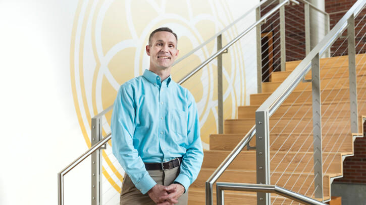 David Ward stands on the stairs of the PLU nursing center. There is a large gold image of the PLU rose window on the wall behind him. He is smiling and wearing a light blue shirt.