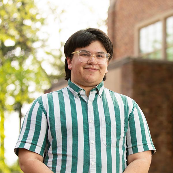 Dylan Ruggeri '23. He is wearing a striped green shirt The photo is taken outside in front of a building on campus.