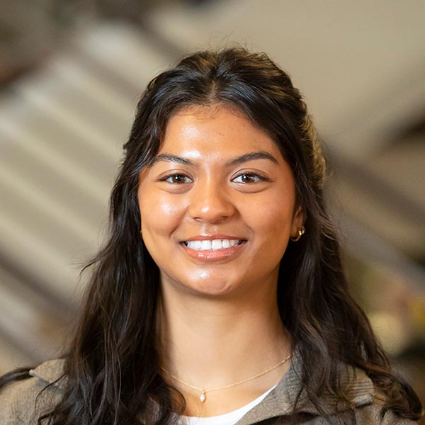 Grayson Nottage stands beneath the staircase in PLU's Anderson University Center. She is smiling and wearing a light brown sweatshirt.