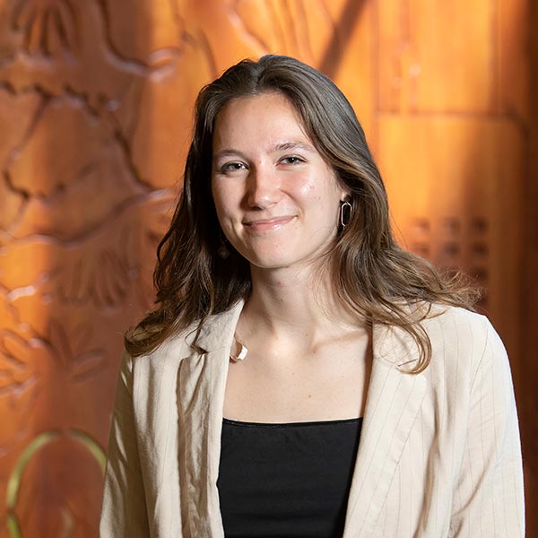 PLU student, wearing a black shirt, cream cardigan sweater, and orange and yellow pants, smiles into the camera while standing in front of a wooden wall and window.